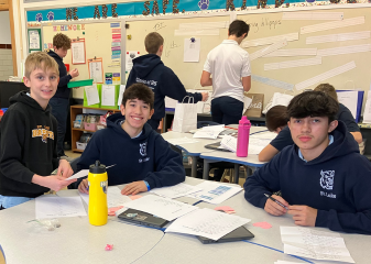 3 boys facing the camera and writing at a table
